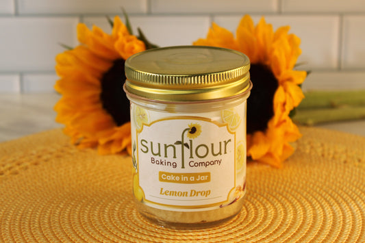 A Sunflour Baking Company Lemon Drop Cake in a Jar sits on a yellow mat, sunflowers in the background.