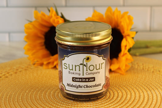 A jar of Sunflour Baking Company Midnight Chocolate Cake in a Jar rests on a woven mat, sunflowers in the background.