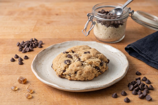 The VGF Classic Chocolate Chip Cookie by Sunflour Baking Company sits on a plate with chocolate chips and sugar, on a wooden surface.