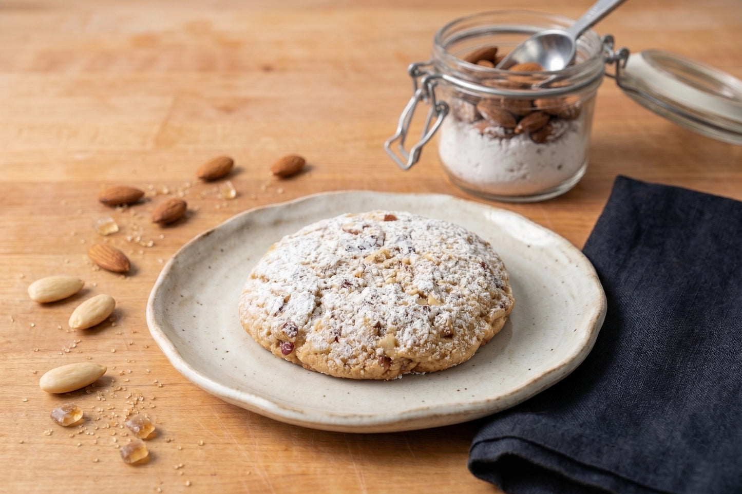 A Swiss Almond Cookie by Sunflour Baking Company on a ceramic plate, with almonds and sugar on a wooden surface.
