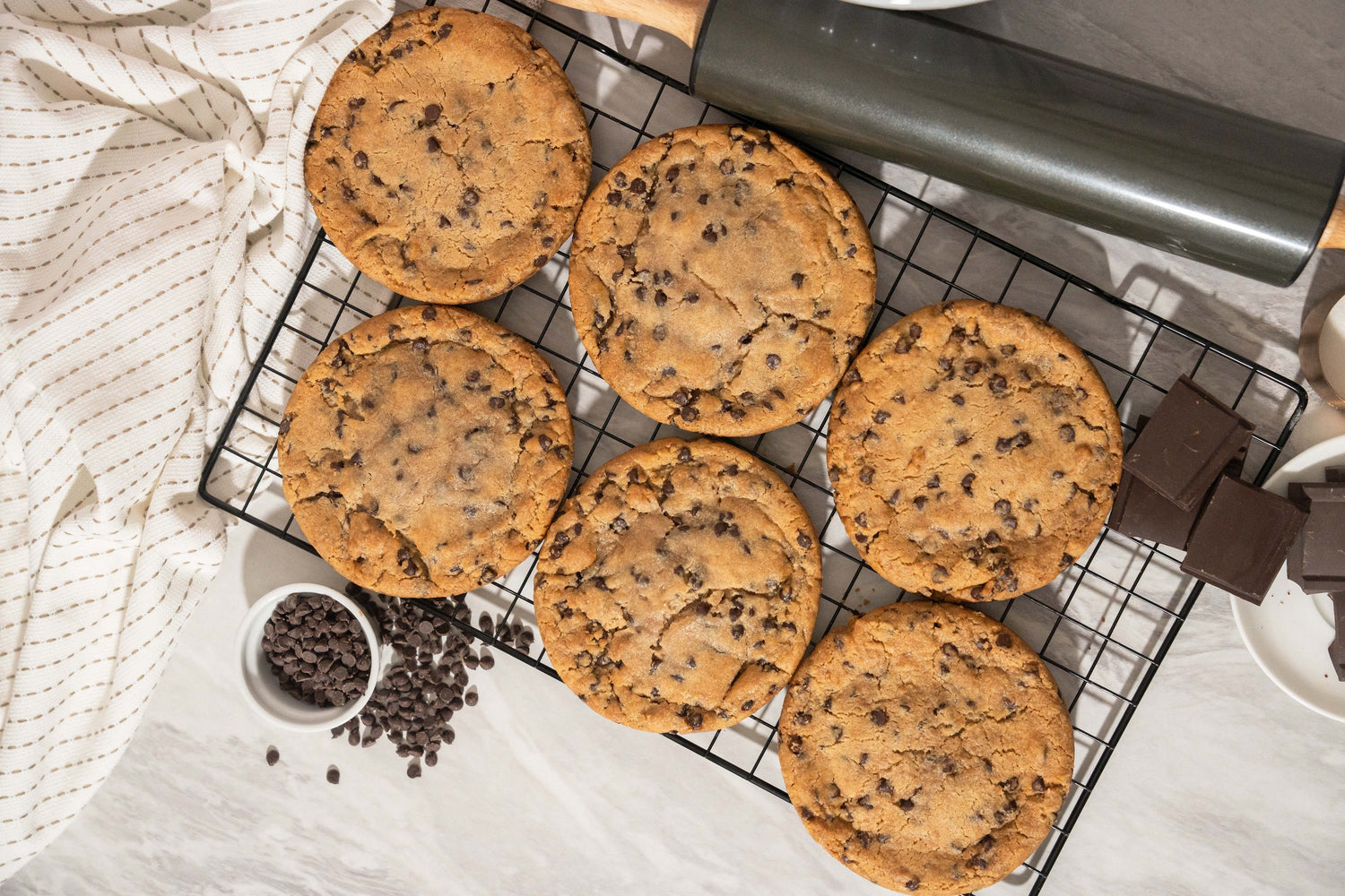 Six Colossal Cookies from the Sunflour Baking Company rest on a cooling rack, surrounded by a rolling pin, chocolate pieces, and a small bowl of chocolate chips. Set on a light-colored surface with a striped cloth, these cookies are truly an irresistible sight for any sweet tooth.
