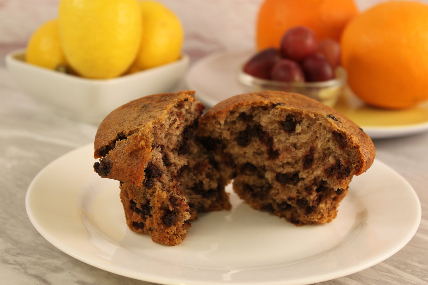 A banana muffin made with Sunflour Baking Company mix, halved on a white plate, with lemons, oranges, and grapes behind it.