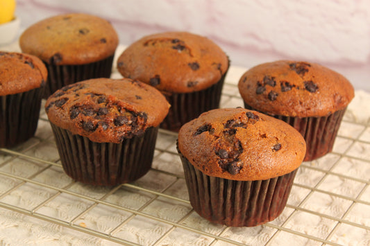 Six Banana Chocolate Muffins by Sunflour Baking Company are cooling on a wire rack set on a textured cloth surface.