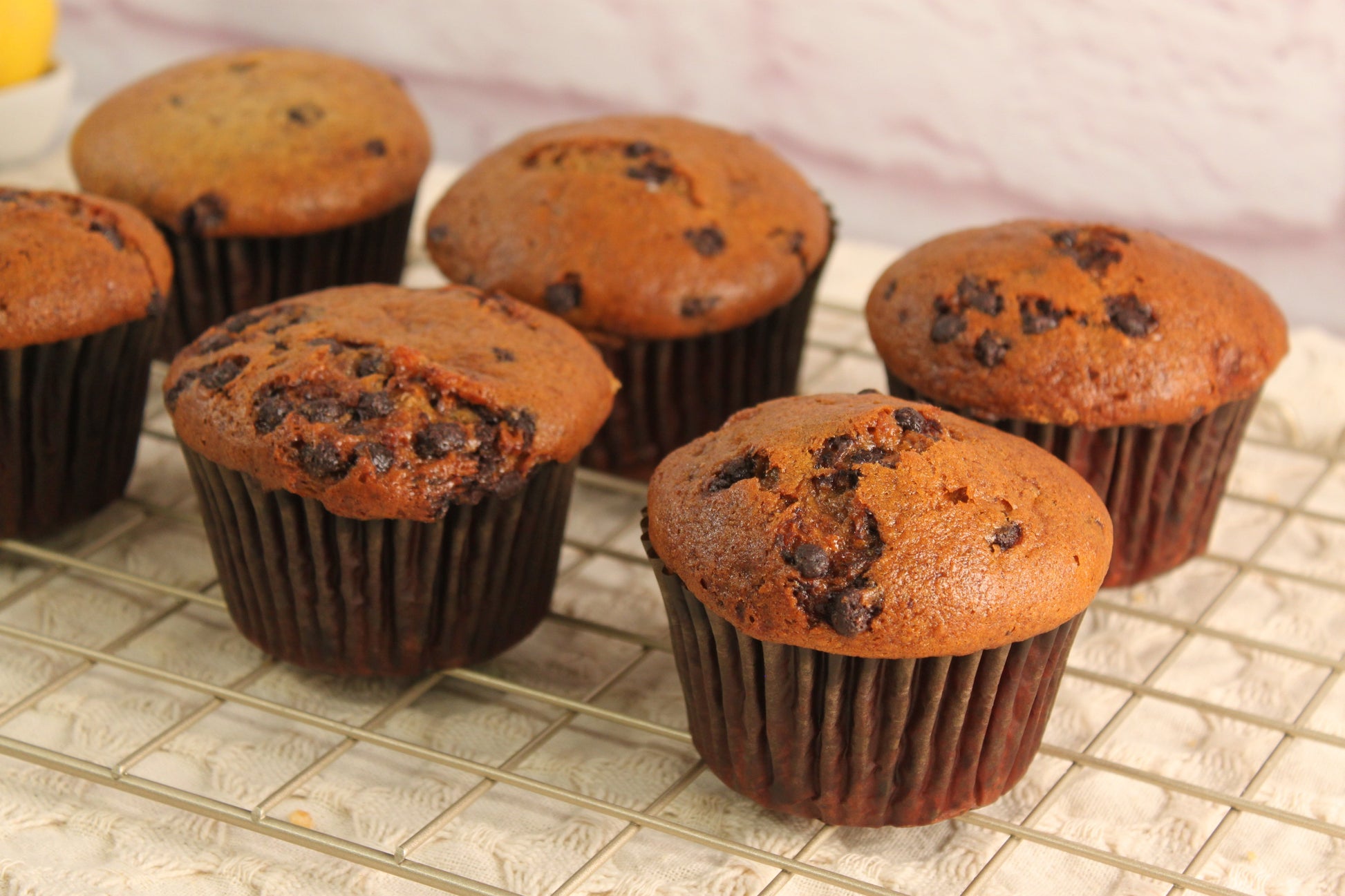 Six Banana Chocolate Muffins by Sunflour Baking Company are cooling on a wire rack set on a textured cloth surface.