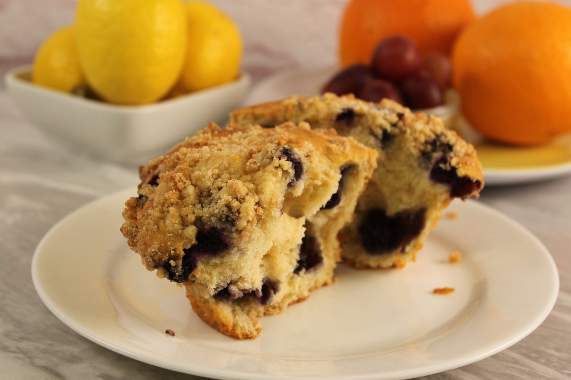 A blueberry muffin made from Sunflour Baking Company Blueberry Muffin Mix is halved on a plate with fruit in the background.