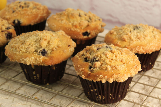 Six Blueberry Muffins from Sunflour Baking Company cooling on a rack, each topped with crumbly streusel.