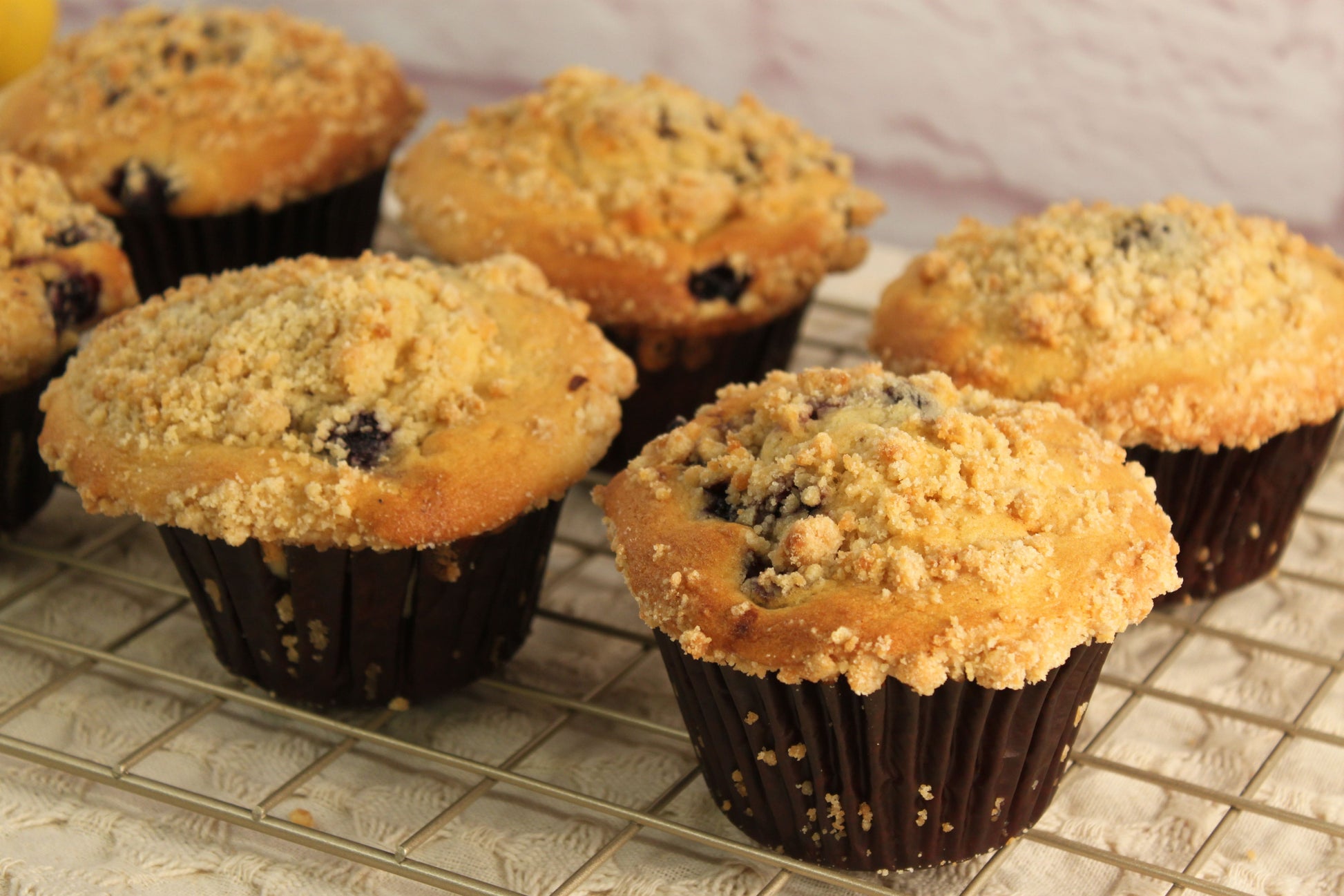 Six Blueberry Muffins from Sunflour Baking Company cooling on a rack, each topped with crumbly streusel.