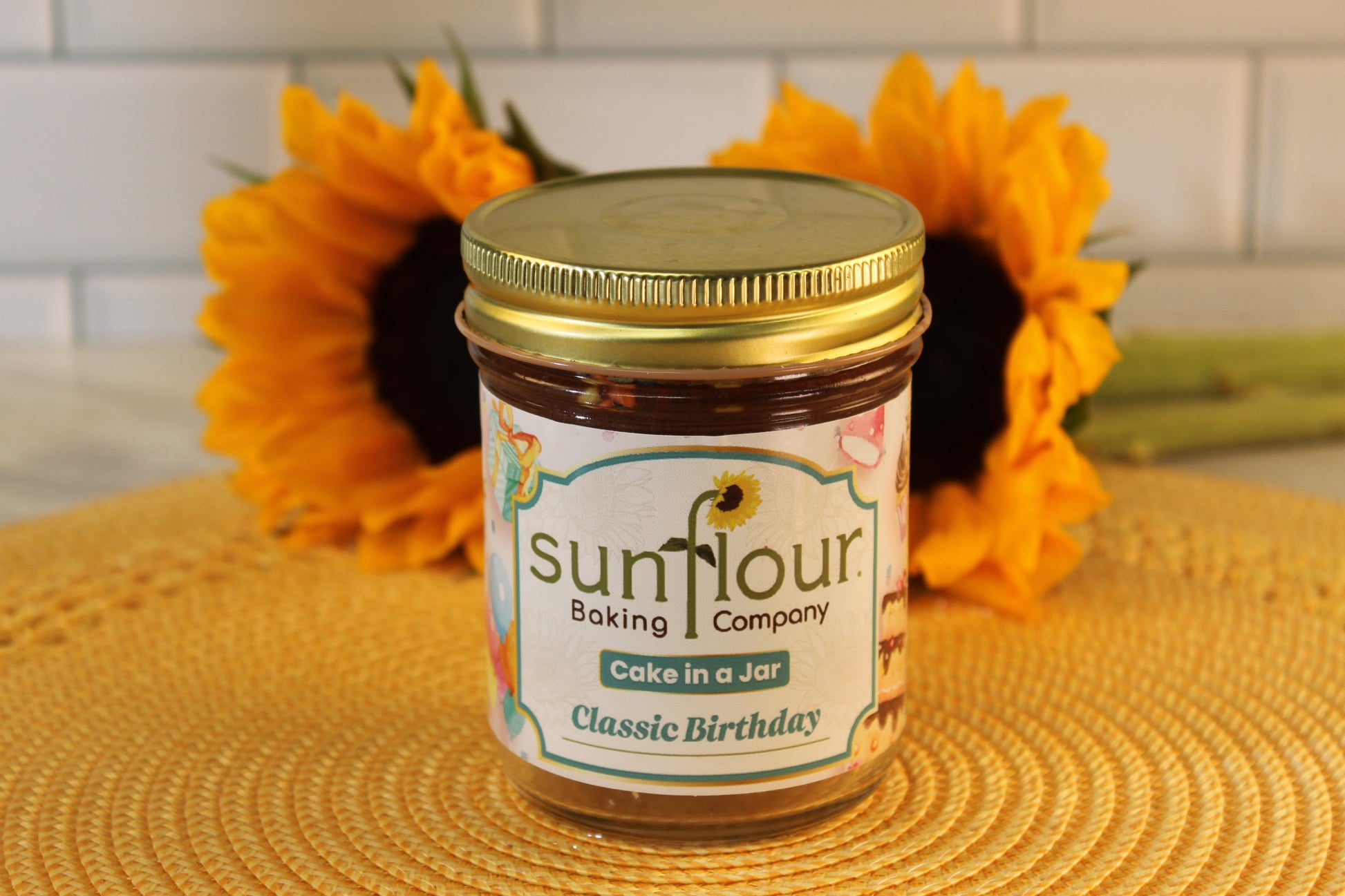 A jar of Sunflour Baking Company Classic Birthday sits on a yellow mat with two sunflowers in the background.