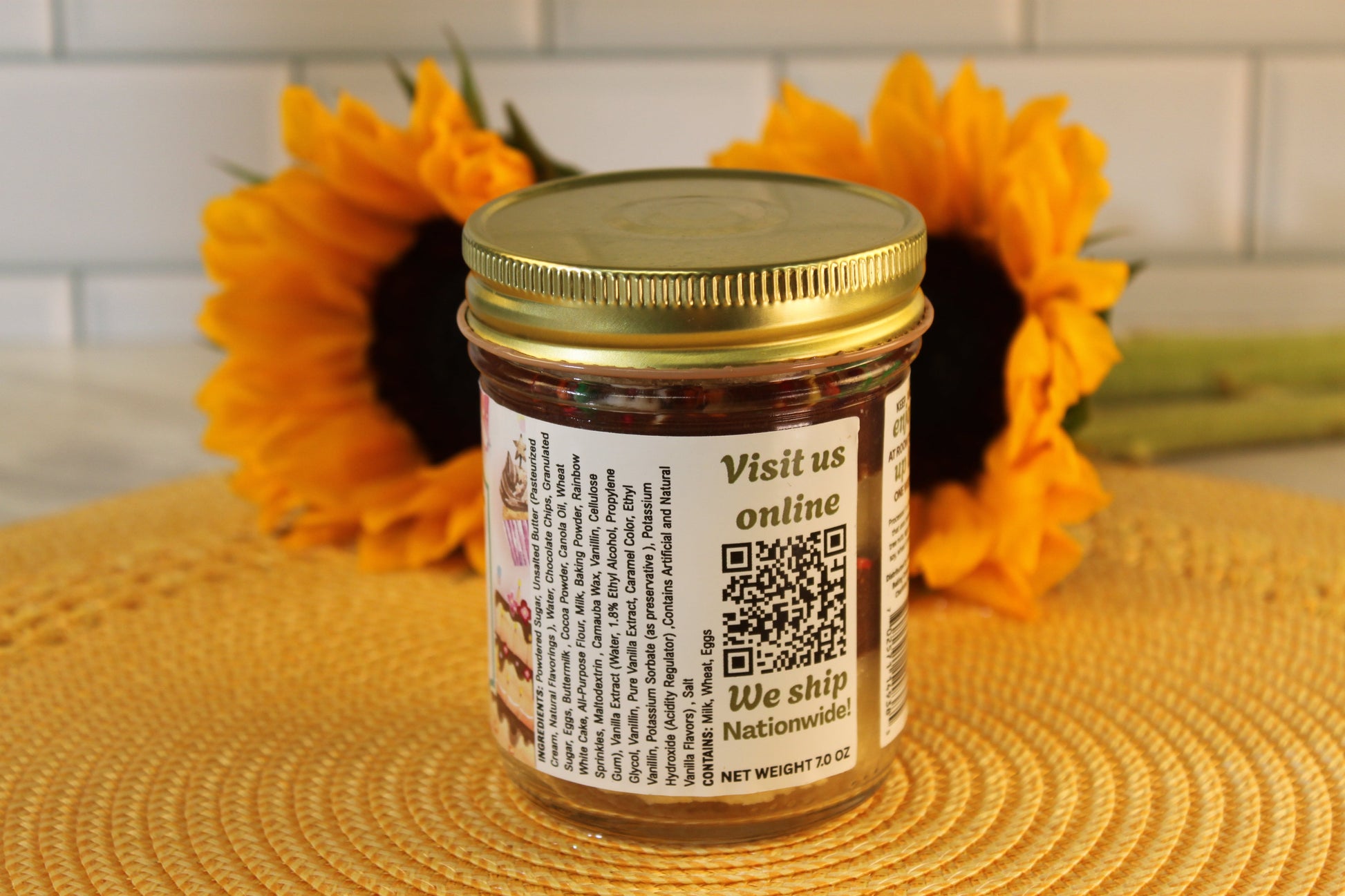 A Classic Birthday jar by Sunflour Baking Company with a gold lid and QR code sits on a mat, evoking cake and chocolate icing.