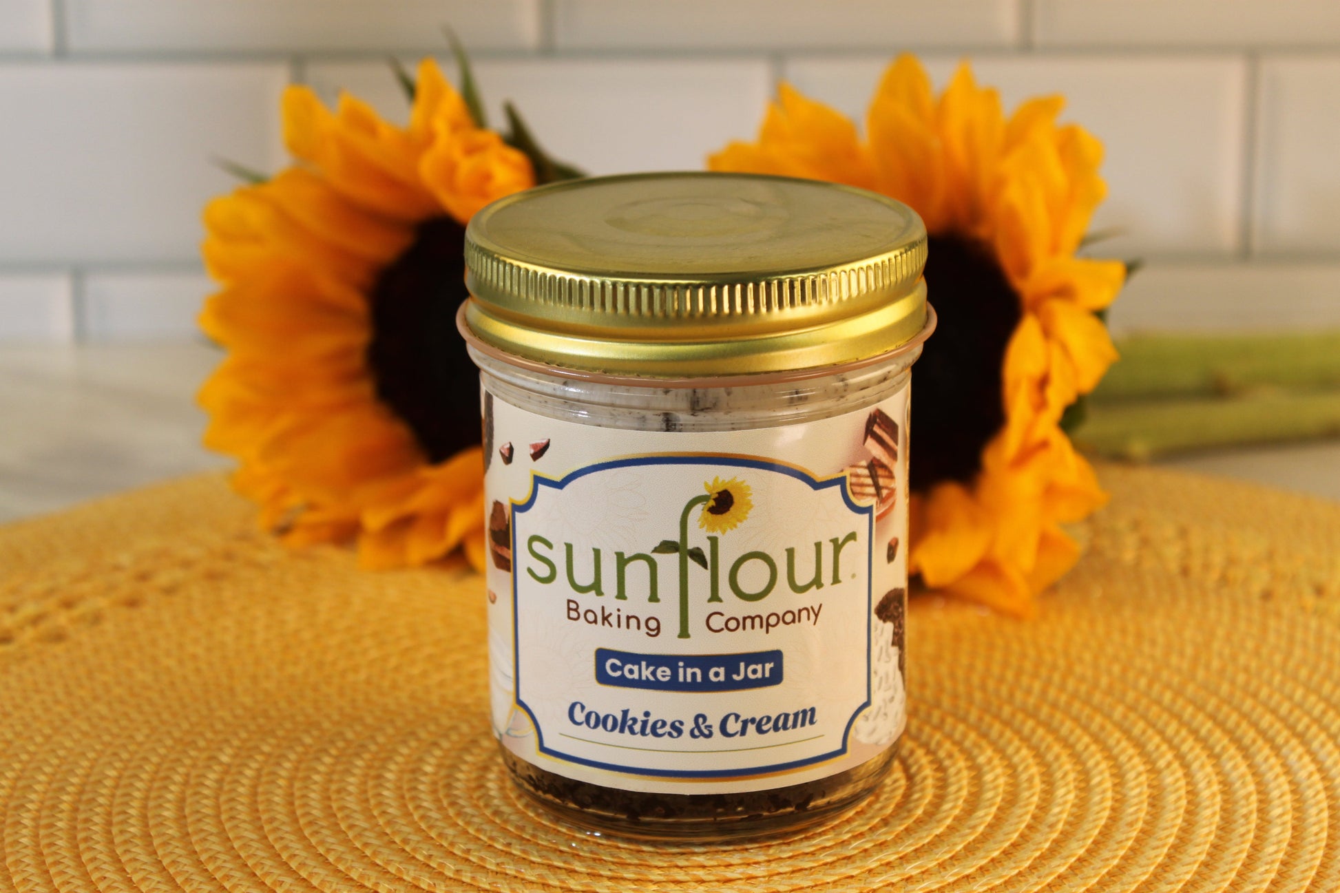 A Sunflour Baking Company Cookies and Cream Cake Jar sits on a yellow mat with sunflowers in the background.