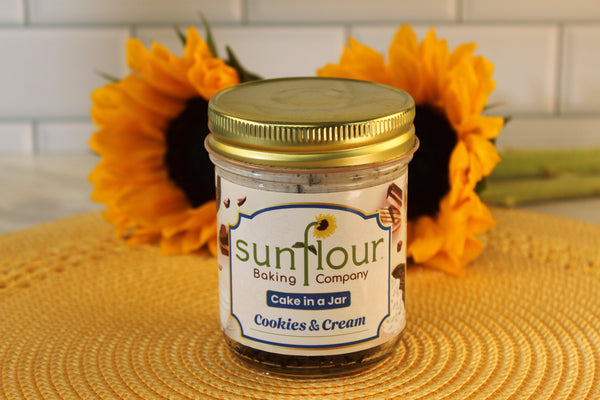 A Sunflour Baking Company Cookies and Cream Cake Jar sits on a yellow mat with sunflowers in the background.