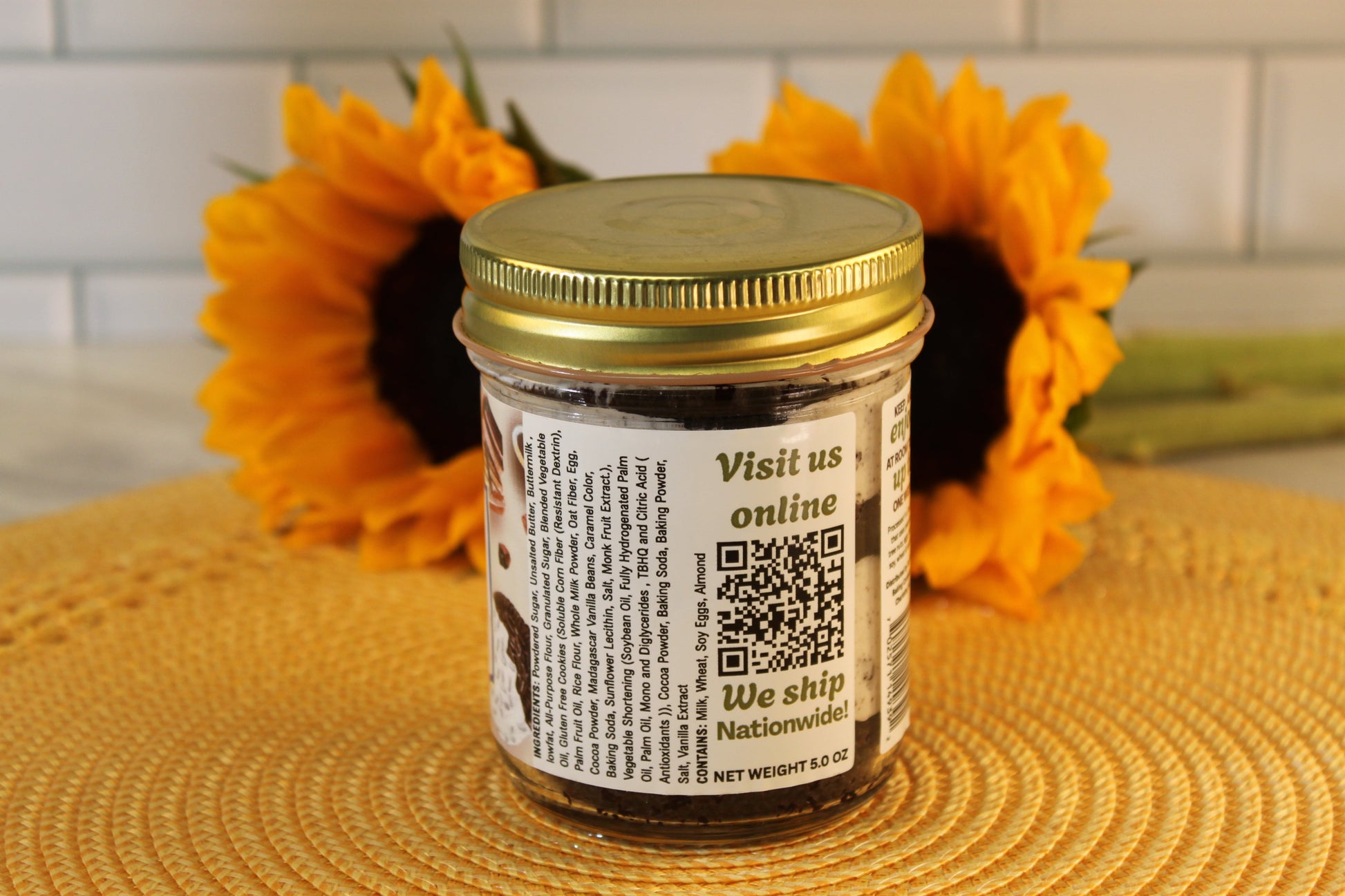 A glass jar of Sunflour Baking Company’s Cookies and Cream, with a gold lid, label QR code, sunflowers, and cookie pieces behind.