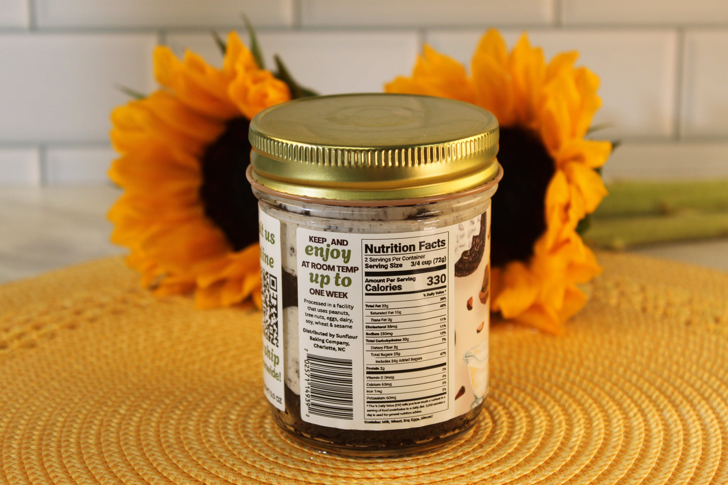 A glass jar of Sunflour Baking Company Cookies and Cream with a gold lid sits on a mat, in front of two sunflowers.