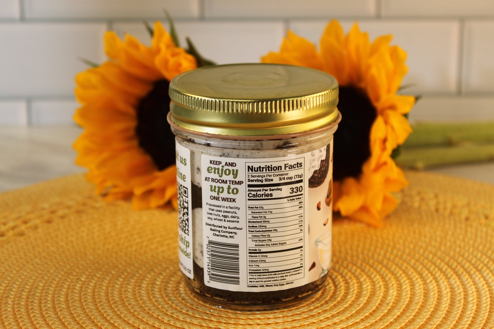 A glass jar of Sunflour Baking Company Cookies and Cream with a gold lid sits on a mat, in front of two sunflowers.