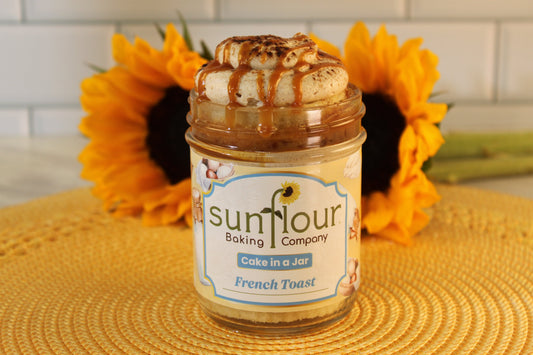 A Sunflour Baking Company French Toast Cake in a Jar sits on a yellow mat with two sunflowers behind it.