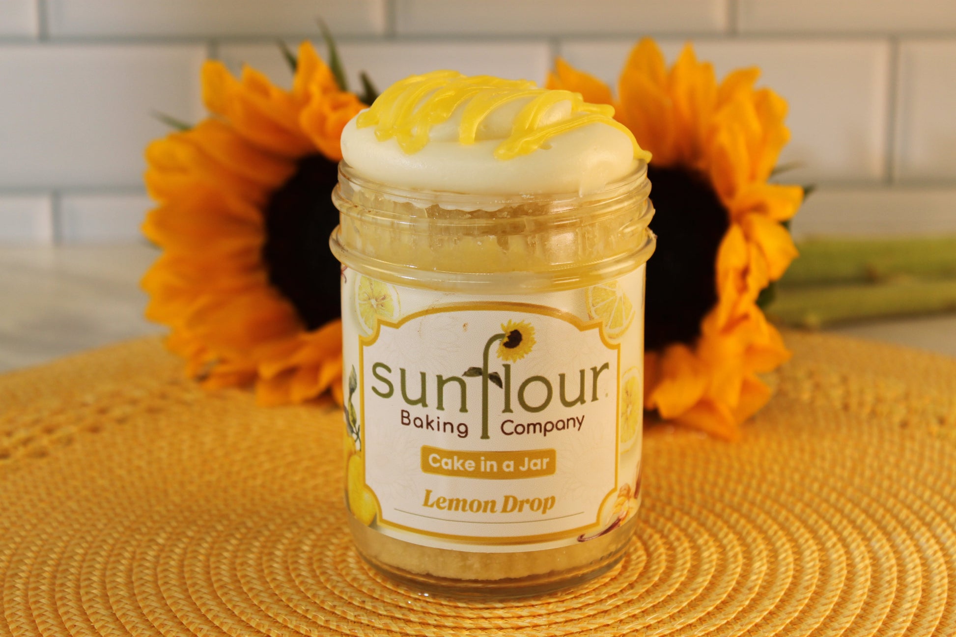 A Sunflour Baking Company Lemon Drop jar sits on a woven mat with sunflowers and a white tile background.