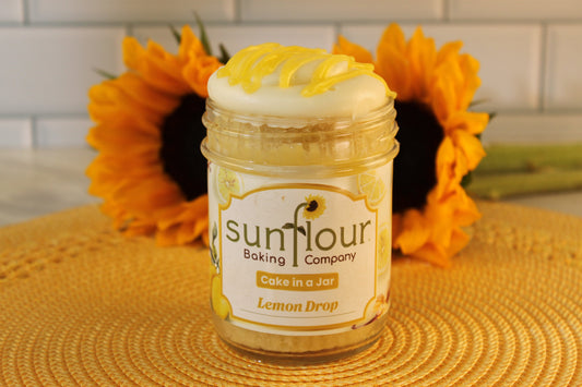 A Sunflour Baking Company Lemon Drop jar sits on a woven mat with sunflowers and a white tile background.