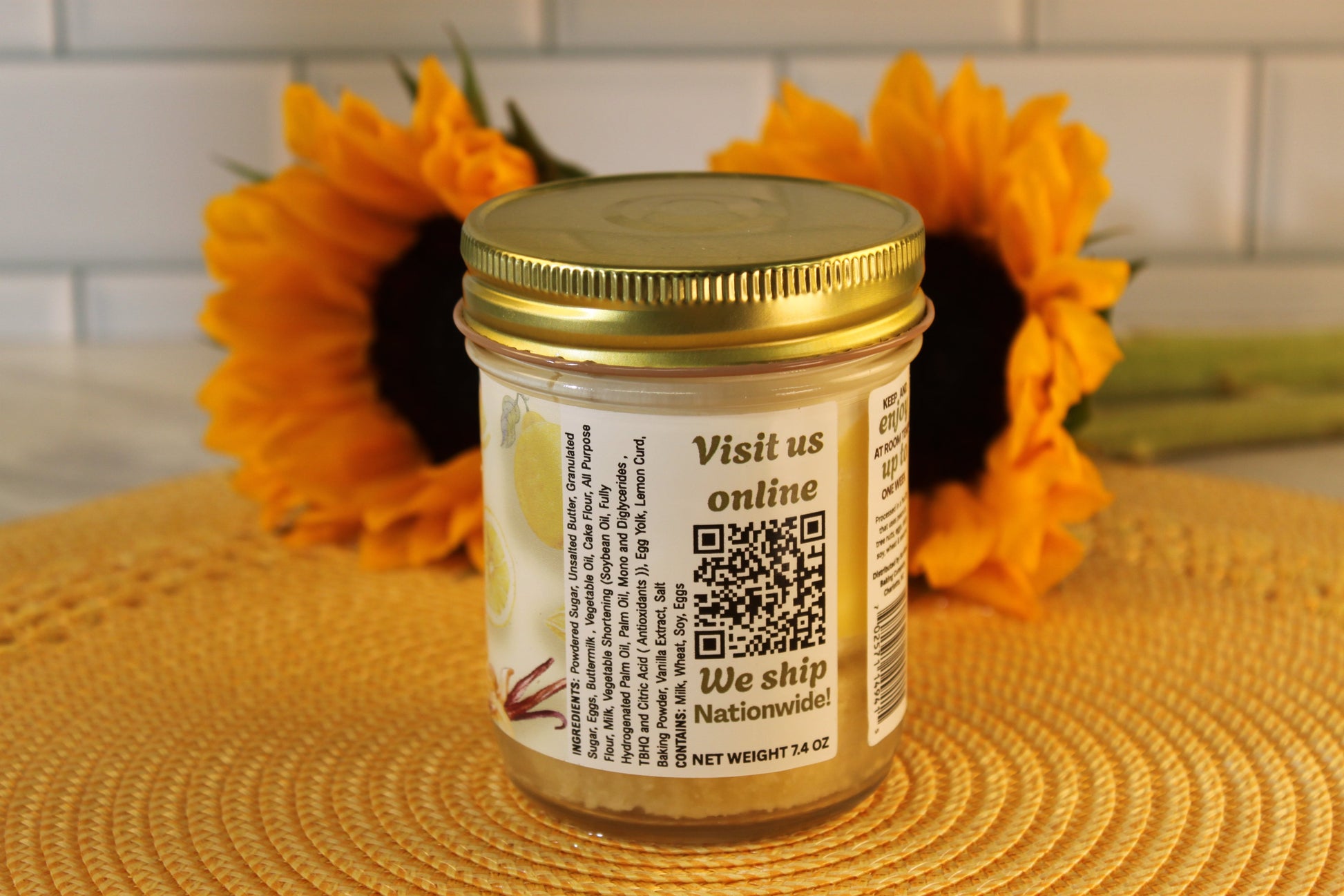 A jar of Lemon Drop by Sunflour Baking Company with a gold lid and QR code sits on a woven mat, sunflowers behind.