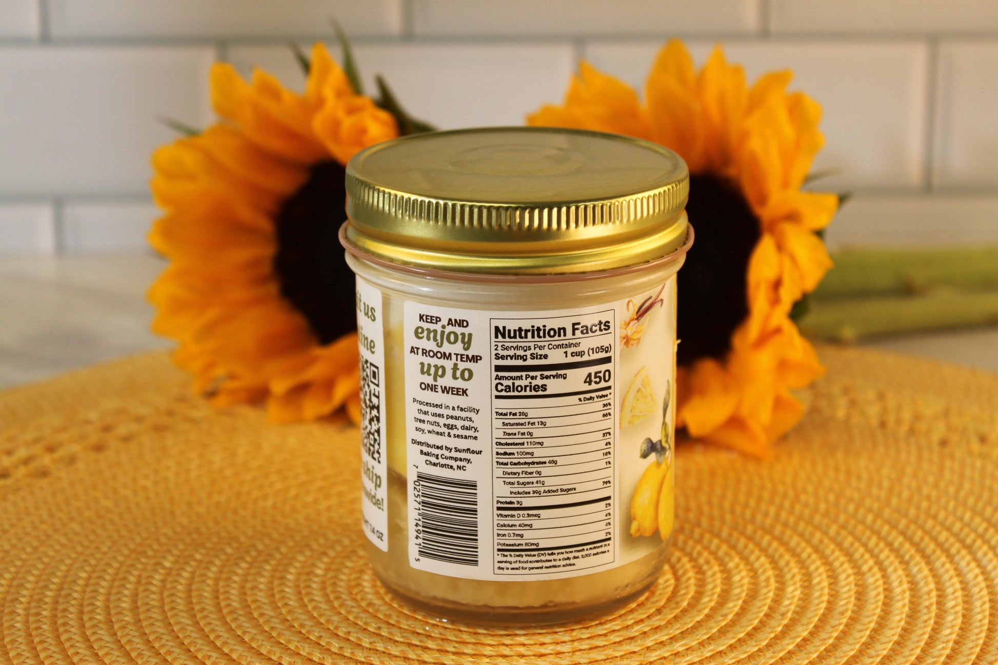 A Sunflour Baking Company Lemon Drop jar with a gold lid shows nutrition facts, set on a placemat with two sunflowers behind.