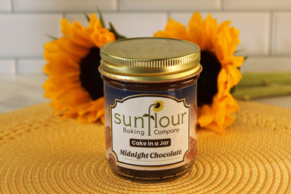 A Midnight Chocolate Cake Jar by Sunflour Baking Company rests on a woven mat with sunflowers—an ideal dessert gift.