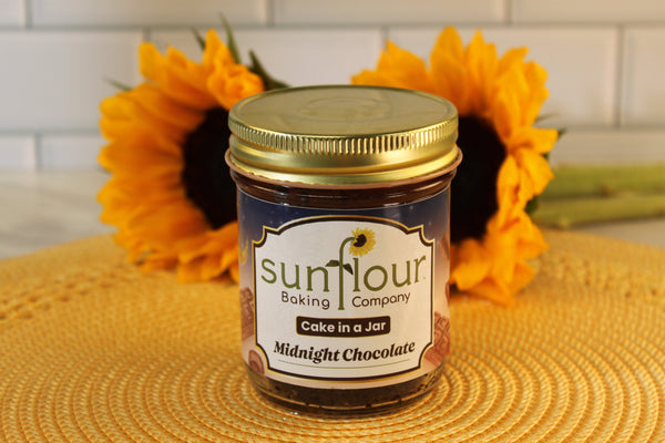 A Midnight Chocolate Cake Jar by Sunflour Baking Company rests on a woven mat with sunflowers—an ideal dessert gift.