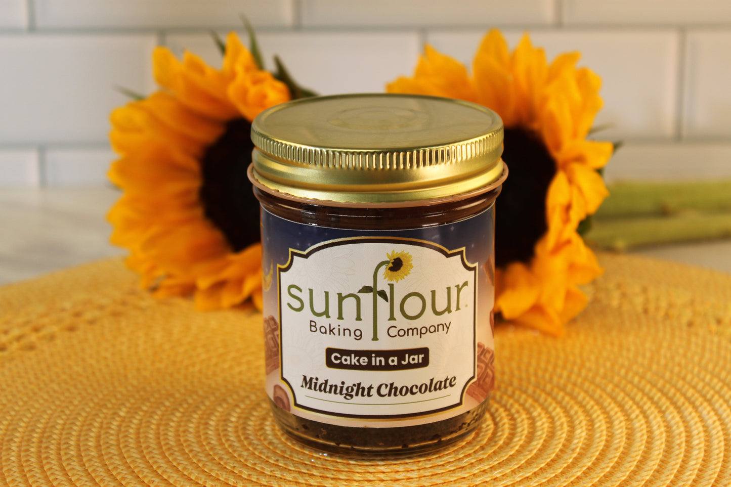 A jar of Sunflour Baking Company Midnight Chocolate Cake in a Jar rests on a woven mat, sunflowers in the background.