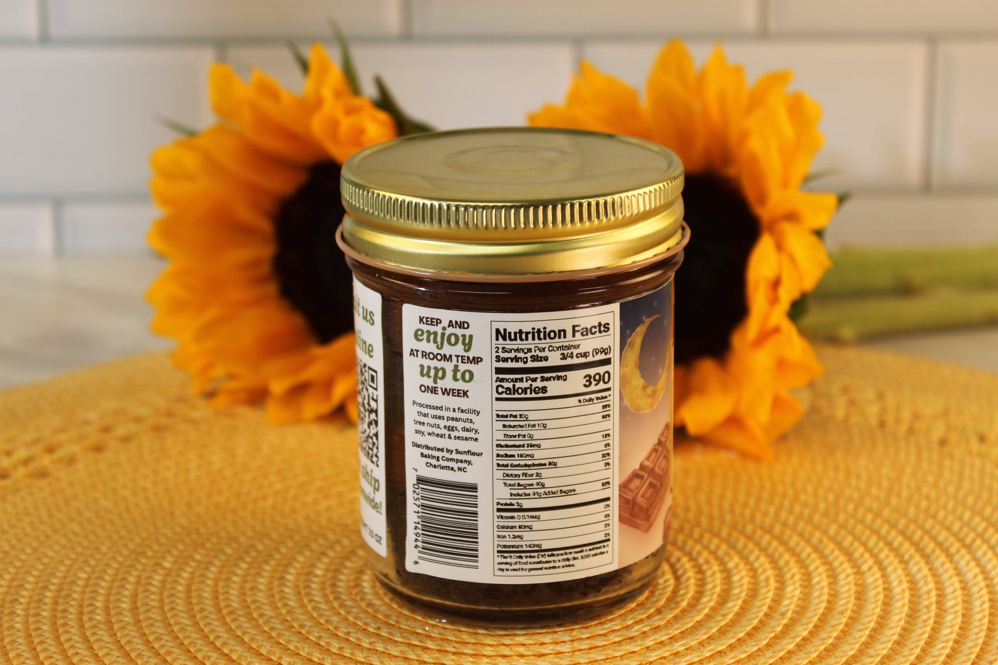A jar of Sunflour Baking Company’s Midnight Chocolate with a gold lid, nutrition label, and two sunflowers on a kitchen counter.