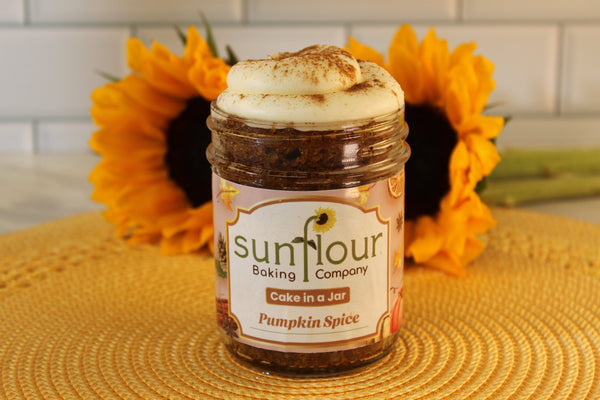 A Sunflour Baking Company Pumpkin Spice Cake Jar with frosting sits on a woven mat, sunflowers in the background.