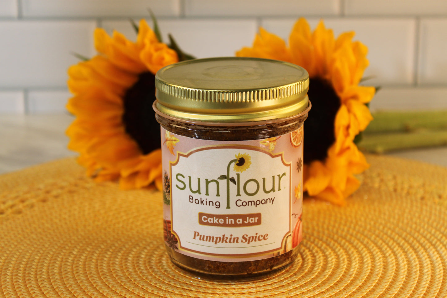 A Pumpkin Spice Cake Jar by Sunflour Baking Company sits on a woven mat with sunflowers in the background.