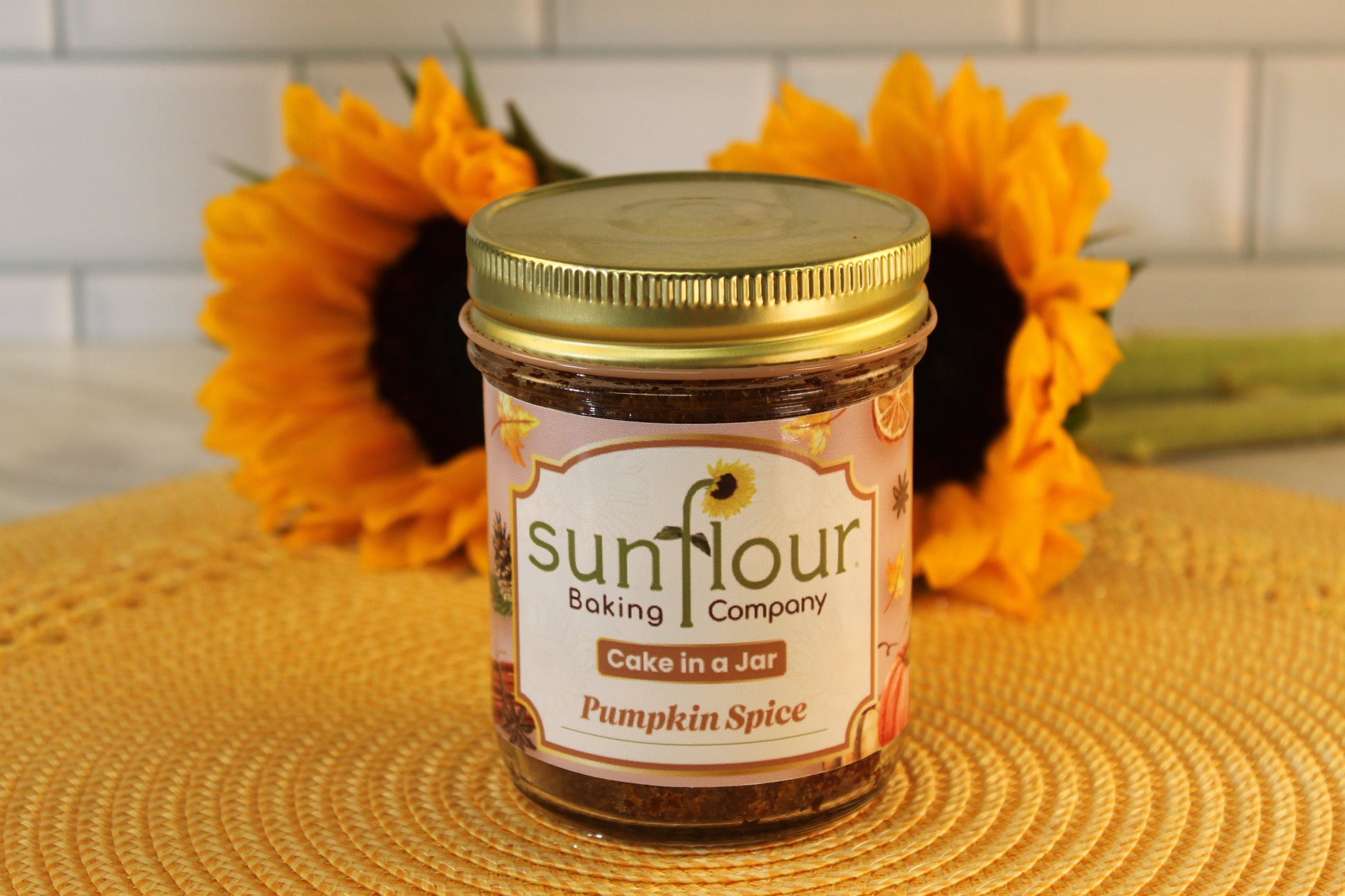 A Pumpkin Spice Cake Jar by Sunflour Baking Company sits on a woven mat with sunflowers in the background.