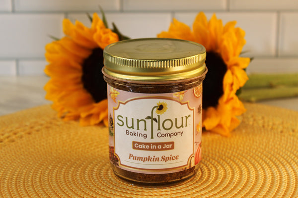 A Pumpkin Spice Cake Jar by Sunflour Baking Company sits on a woven mat with sunflowers in the background.