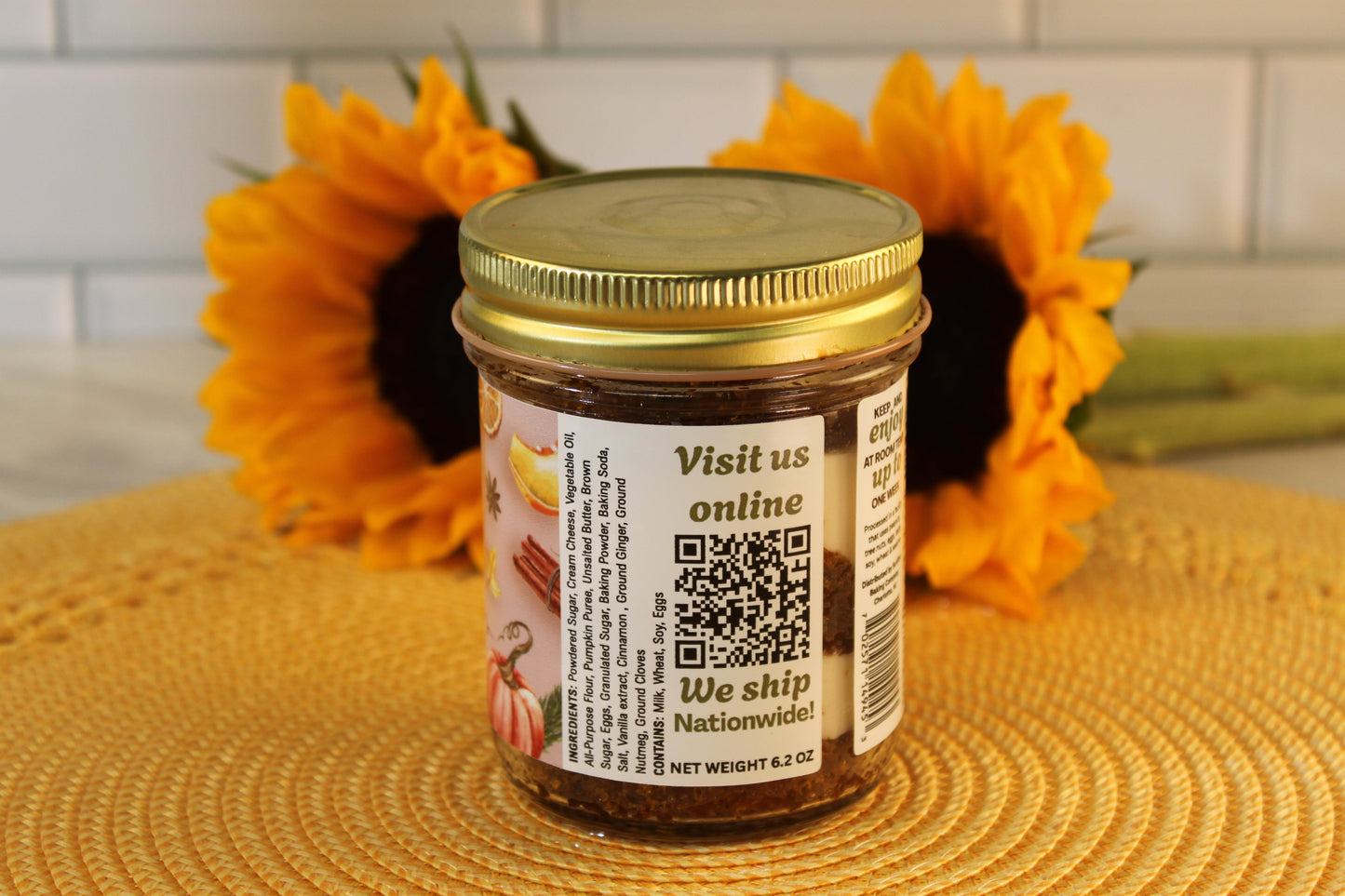 A Sunflour Baking Company Pumpkin Spice jar with gold lid and QR label sits on a yellow placemat, sunflowers in the background.