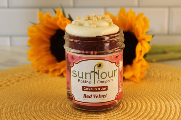 A Sunflour Baking Company Red Velvet Cake Jar with cream cheese frosting sits on a yellow mat, sunflowers in the background.