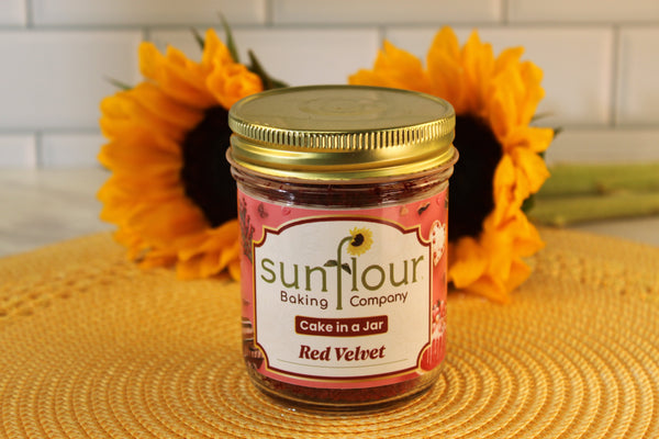 A Sunflour Baking Company Red Velvet Cake Jar with cream cheese frosting is on a yellow mat, sunflowers in the background.