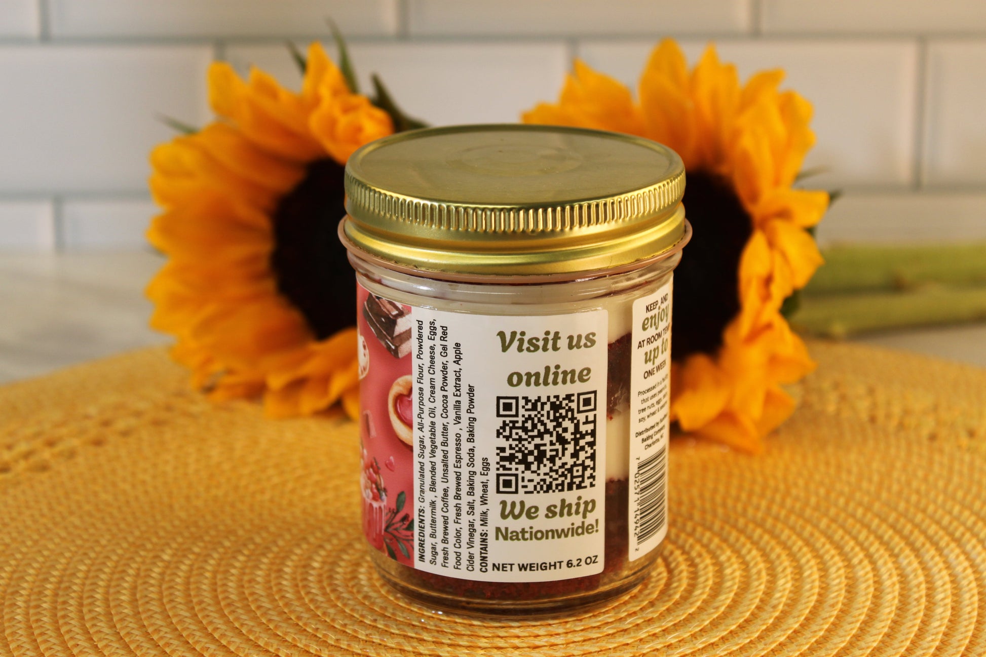A Red Velvet Cake Jar by Sunflour Baking Company with a gold lid sits on a mat, featuring cream cheese frosting and a QR code label.