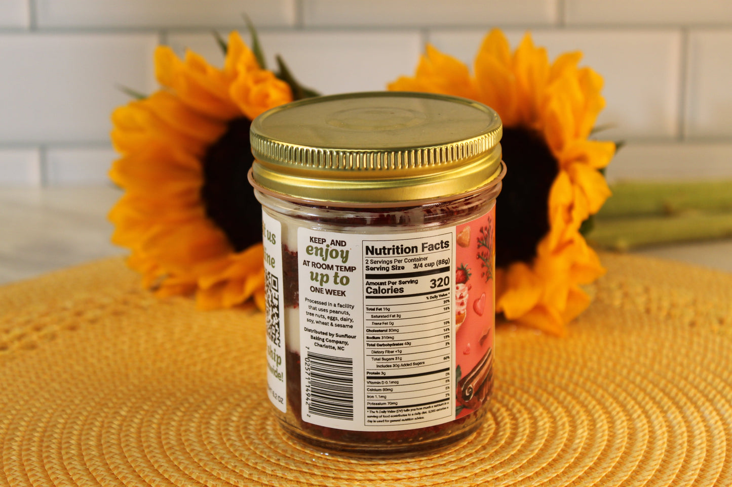 A Red Velvet jar from Sunflour Baking Company with a gold lid sits on a placemat; sunflowers are in the background.