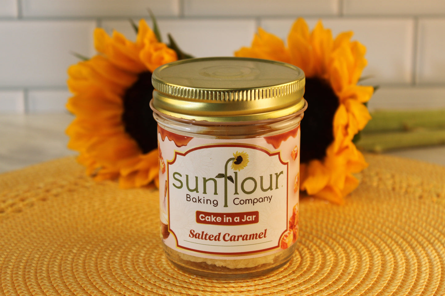 A Sunflour Baking Company Salted Caramel cake jar, pictured with sunflowers and a woven mat in the background.