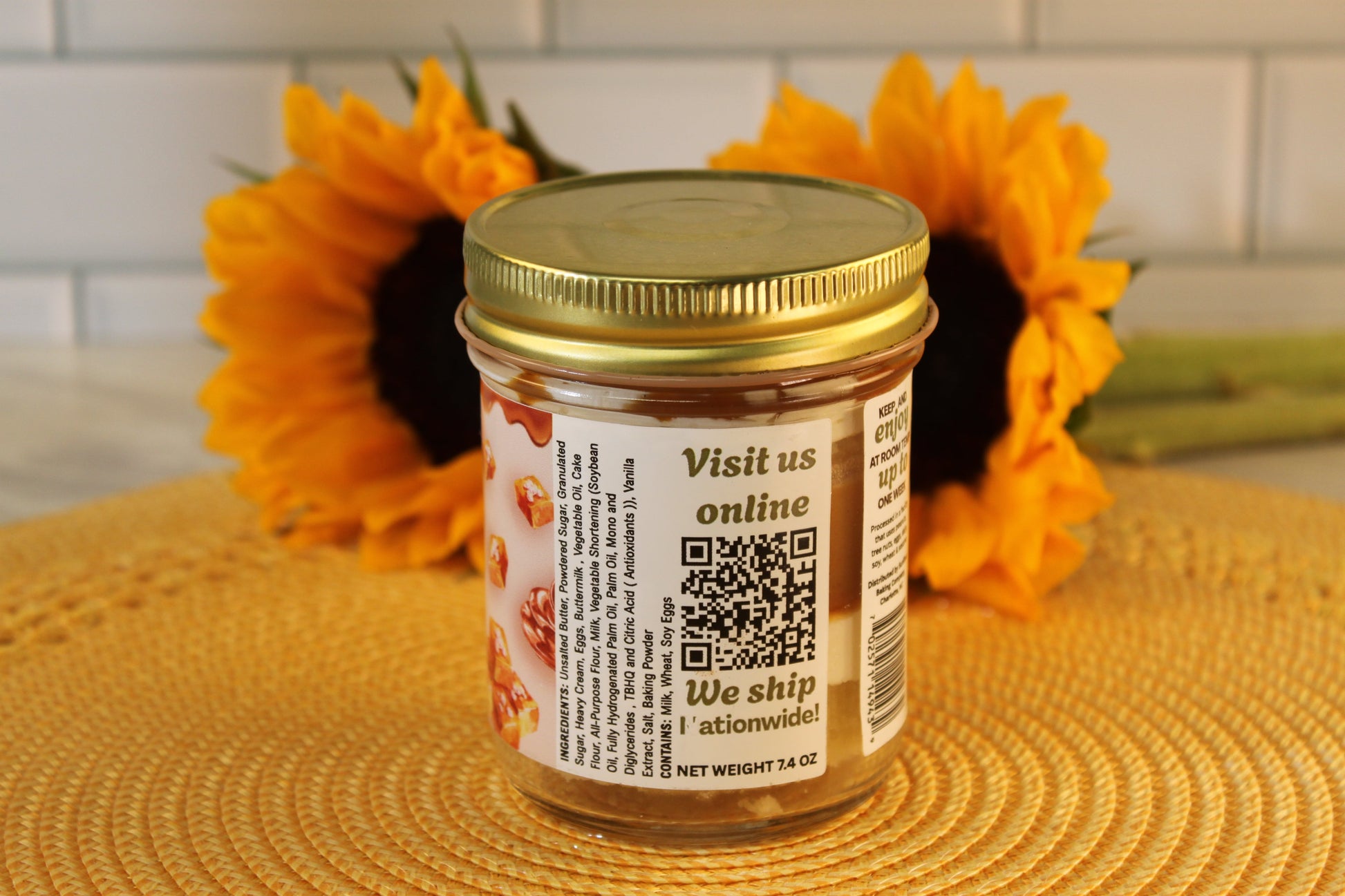A Salted Caramel jar by Sunflour Baking Company with a gold lid sits on a yellow mat, labeled with a QR code and sunflowers behind.