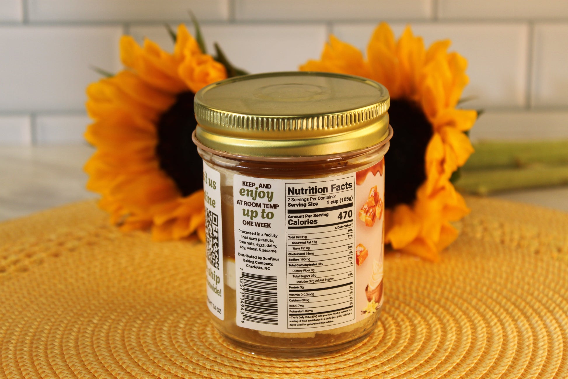 A Salted Caramel jar by Sunflour Baking Company with a gold lid sits on a mat, two sunflowers in the background.