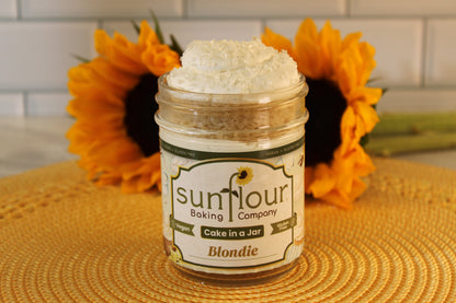 A Sunflour Baking Company VGF Blondie Cake Jar with frosting sits on a woven mat, flanked by sunflowers in the background.