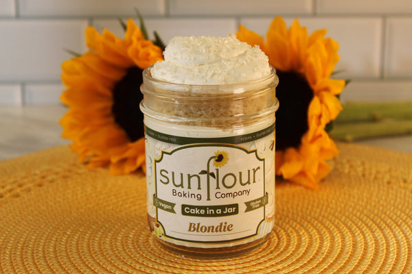 A Sunflour Baking Company VGF Blondie Cake Jar with frosting sits on a woven mat, flanked by sunflowers in the background.