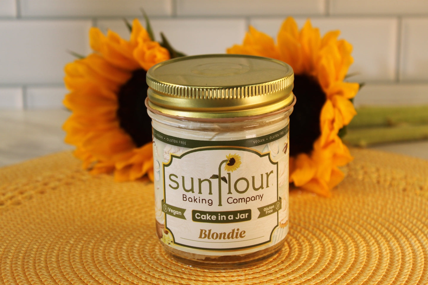 A Sunflour Baking Company VGF Blondie Cake Jar sits on a yellow mat with two sunflowers, featuring gluten-free cake.