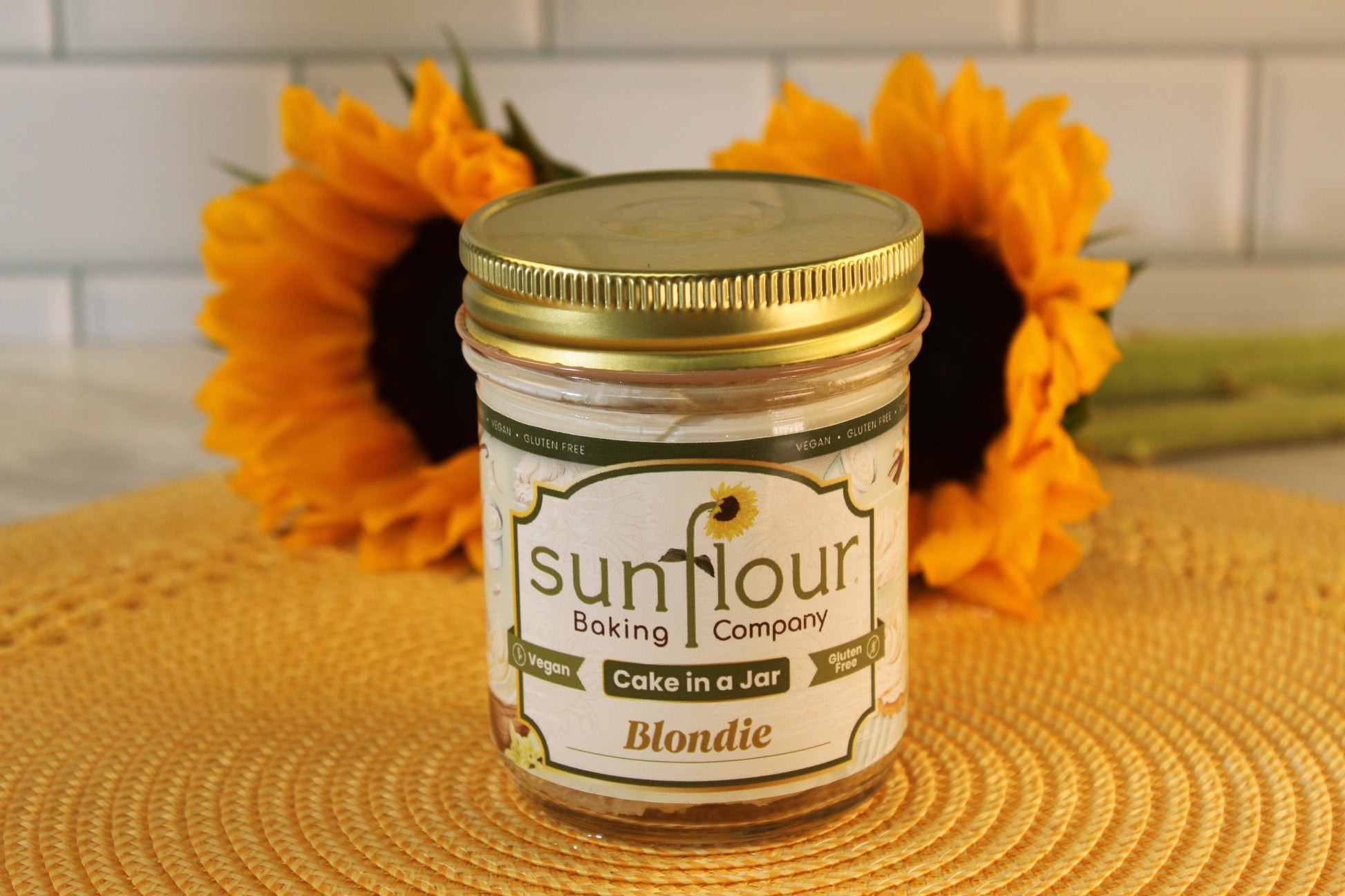 A Sunflour Baking Company VGF Blondie Cake Jar sits on a yellow mat with two sunflowers, featuring gluten-free cake.
