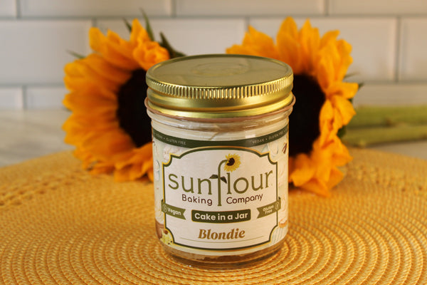A Sunflour Baking Company VGF Blondie Cake Jar sits on a yellow mat with two sunflowers, featuring gluten-free cake.