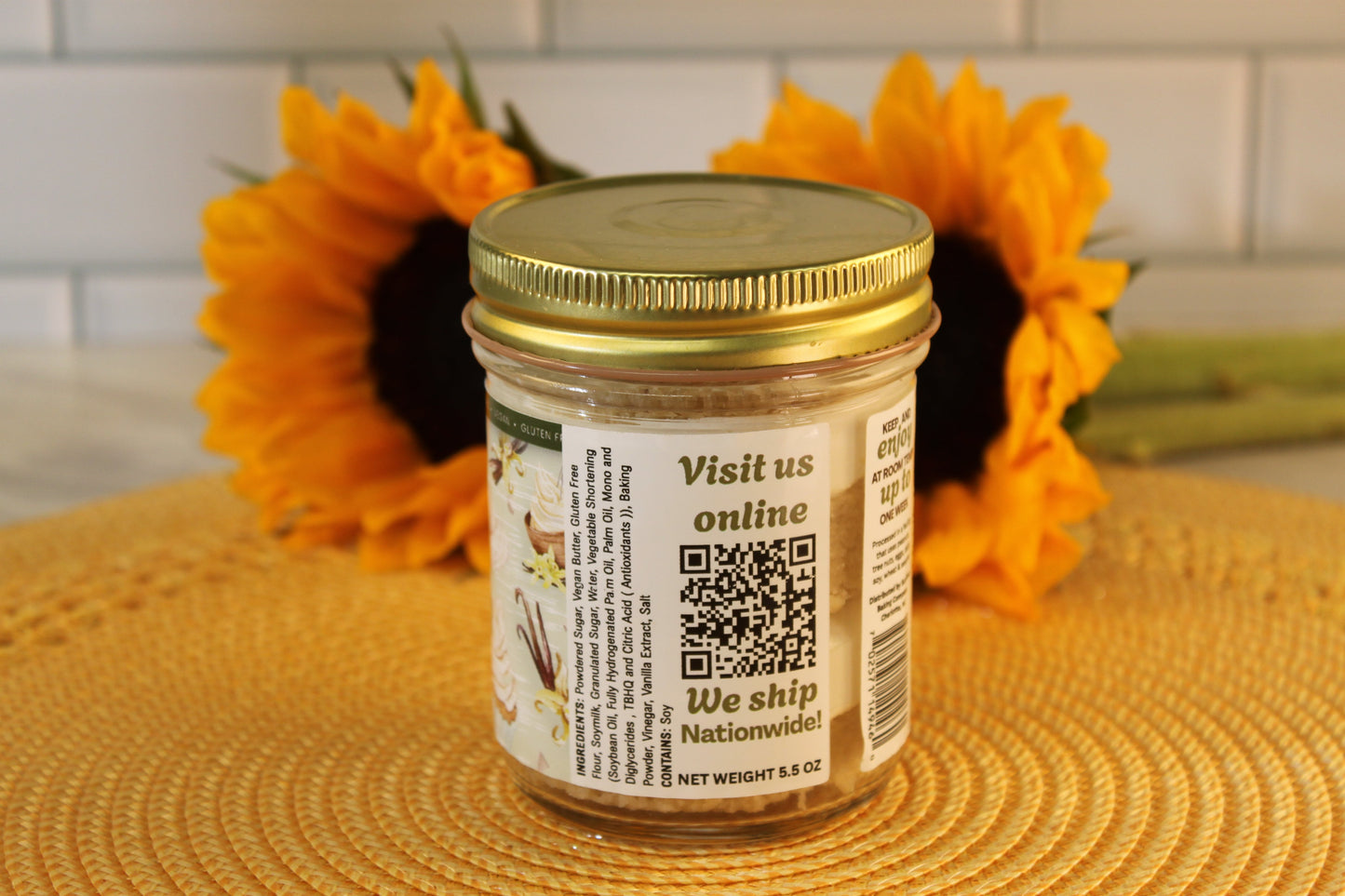 A VGF Blondie by Sunflour Baking Company in a glass jar with gold lid sits on a yellow placemat with two sunflowers behind.