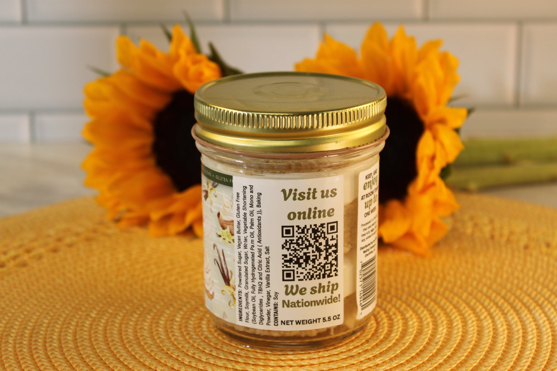 A VGF Blondie by Sunflour Baking Company in a glass jar with gold lid sits on a yellow placemat with two sunflowers behind.