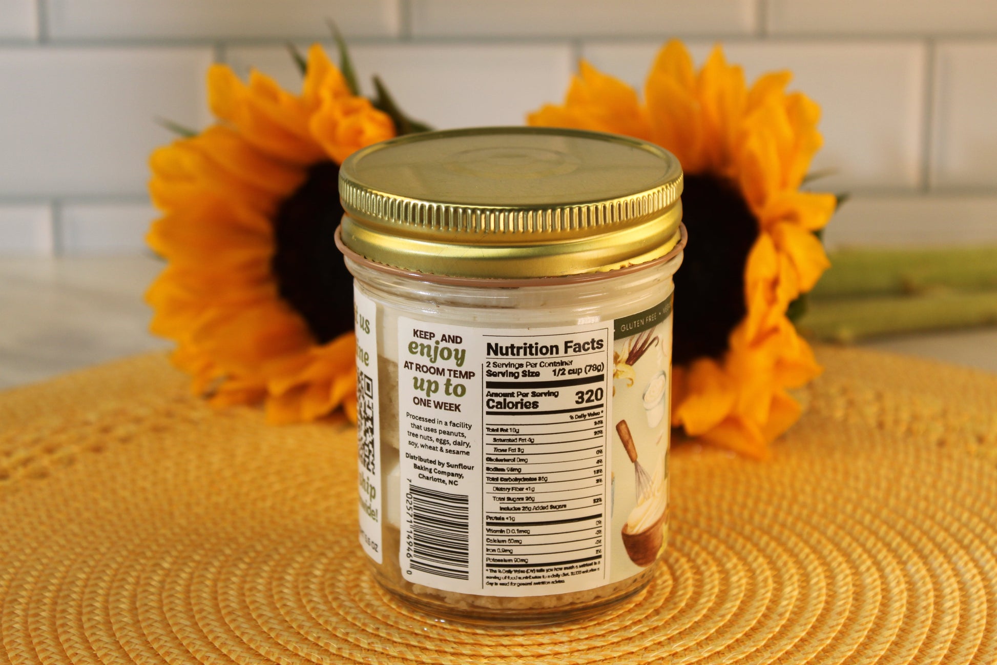 A glass jar of VGF Blondie by Sunflour Baking Company sits on a yellow placemat with two sunflowers in the blurred background.