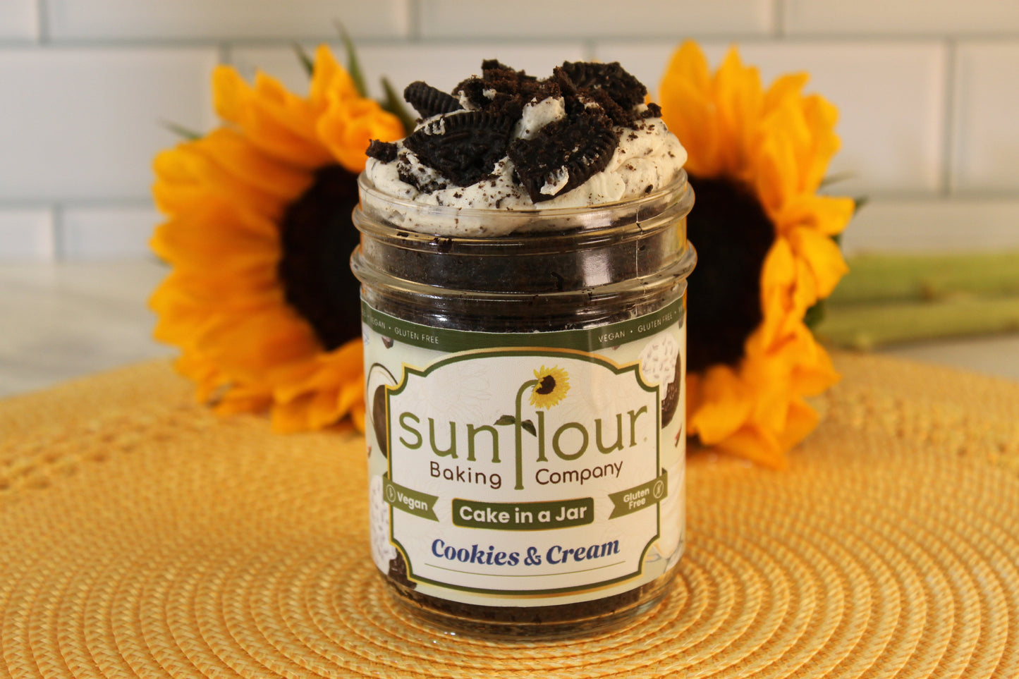 A VGF Cookies and Cream Cake Jar by Sunflour Baking Company rests on a placemat with sunflowers in the background.