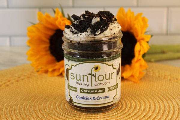 A VGF Cookies and Cream Cake Jar by Sunflour Baking Company rests on a placemat with sunflowers in the background.