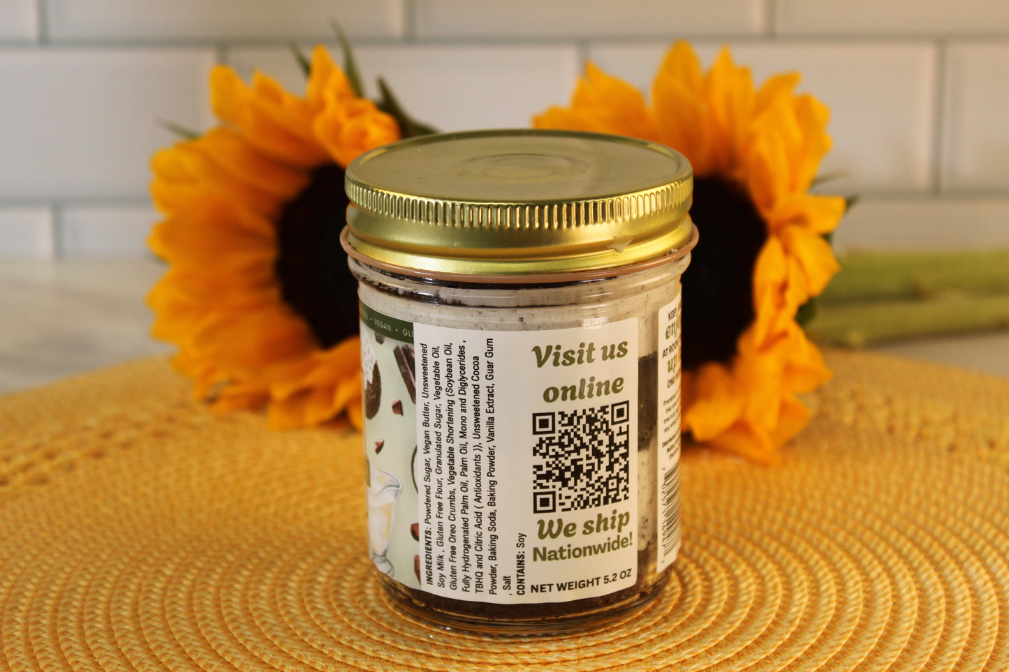 A jar of VGF Cookies and Cream by Sunflour Baking Company with a gold lid sits on a mat; two sunflowers bloom behind it.